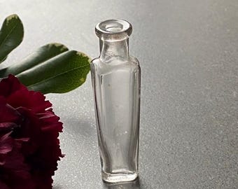 Puede incluir: Una botella de vidrio transparente con forma cuadrada y parte superior redondeada. La botella está vacía y tiene una superficie ligeramente texturizada.