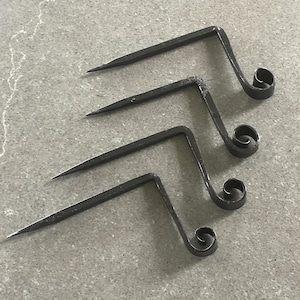 May include: Four handcrafted, dark metal hooks with a sharp point on one end and a decorative scroll on the other. The hooks are arranged on a gray surface, showcasing their unique, forged design.