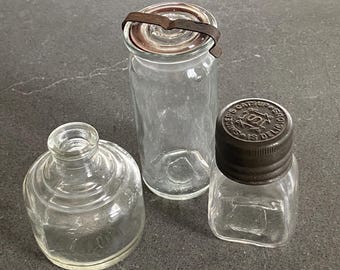 Puede incluir: Tres botellas de vidrio vintage de diferentes formas y tamaños. La botella de la izquierda tiene una boca ancha y un cuerpo redondeado. La botella del centro tiene una forma alta y cilíndrica con una tapa de metal. La botella de la derecha tiene una forma cuadrada y una tapa de metal con el texto "Sanders Catsup Is Delicious".