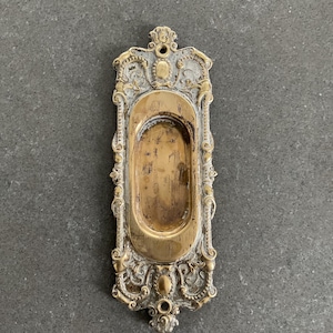 May include: An antique brass door pull with an oval recessed handle. The decorative frame features detailed scrollwork and a distressed, aged appearance. The brass has a warm, golden hue, with hints of silver. A vintage-style hardware piece, suitable for doors.