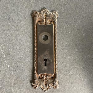 May include: An antique, ornate door plate with a keyhole and a circular hole. The metal plate has a rectangular shape with decorative scrollwork around the edges. The plate has a weathered, aged appearance.