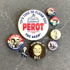 May include: A collection of vintage political campaign buttons featuring various candidates and slogans, including a large white Perot button, a Carter-Mondale button, a William Howard Taft button, two Kennedy buttons, a McKinley and Hobart button, and an elephant pin.