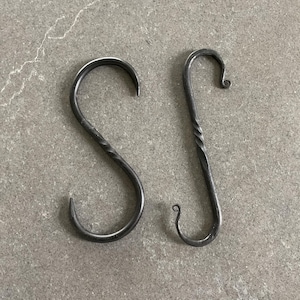 May include: Two handcrafted, dark metal S-hooks. One hook has a simple, curved design, while the other features a twisted stem. These hooks are ideal for hanging items and add a rustic touch to any space.