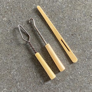 May include: Three vintage knitting tools with ivory-colored handles. The tools are a crochet hook, a yarn needle, and a stitch holder.