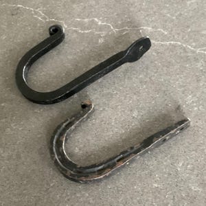 May include: Two wrought iron hooks. One is solid black with a rounded top and a flat end with a hole. The other hook is a horseshoe shape with a distressed finish and multiple holes on the end. Both are on a gray surface.