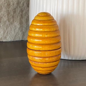 May include: A wooden egg-shaped box with a honeycomb pattern. The box is made of multiple layers of wood, creating a unique and intricate design. The box is painted in a warm, honey-colored hue.