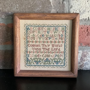 May include: A framed cross-stitch sampler with a wooden frame. The sampler features an alphabet, numbers, and the text "Commit Thy Works Unto The Lord." The embroidery uses green, pink, and blue threads on a beige background.
