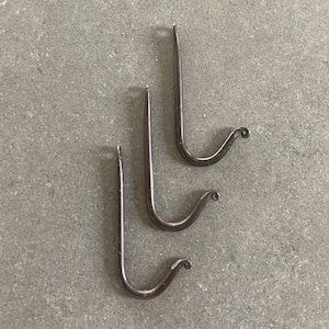 May include: Three handcrafted, dark metal hooks with a curved design. Each hook has a pointed top and a curled end. The hooks are arranged on a gray surface, showcasing their rustic, minimalist aesthetic.
