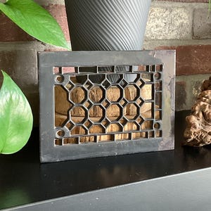 May include: Black metal vent cover with a geometric hexagonal pattern. The vent cover is on a black surface.