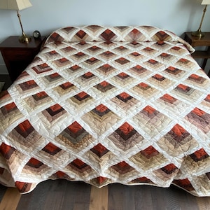 May include: A handmade quilt with a diamond pattern in shades of brown, beige, and rust. The quilt is spread across a bed, showcasing its intricate patchwork design and warm color palette. The quilt's design is a classic, cozy style.