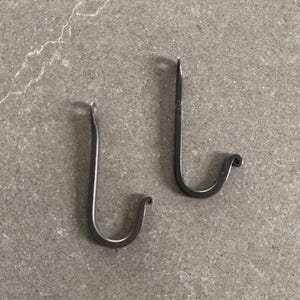 May include: Two handcrafted, dark metal hooks with a curved bottom and a pointed top. The hooks are designed for hanging items and are displayed on a gray surface.