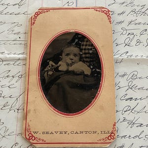 May include: A vintage black and white photograph of a baby in a white dress, sitting in a chair. The photo is framed in a red oval and has a red border. The photo is labeled "W. SEAVEY, CANTON, ILL."