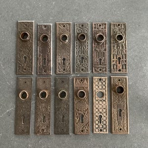 Pode incluir: Uma coleção de doze placas de porta antigas de bronze. Cada placa apresenta um buraco de fechadura e uma abertura circular para um puxador. As placas têm vários padrões em relevo, incluindo desenhos florais, geométricos e de rolagem. Estão dispostas sobre uma superfície cinzenta.