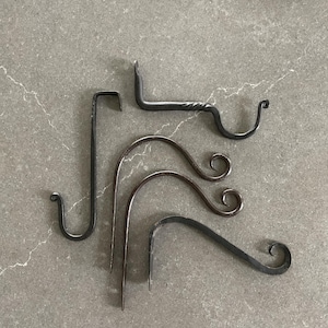 May include: An assortment of handcrafted, dark metal hooks and hangers. The collection includes various shapes, such as a J-hook, a twisted bar hook, and several curved designs with decorative scroll ends. These are likely for hanging plants or decor.