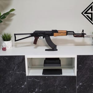 以下が含まれることがあります： 木製のハンドガードとグリップを備えた、黒と茶色のAK-47スタイルのライフルが黒いスタンドに展示されています。ライフルは、白と黒のロゴが付いた白い壁に配置されています。装飾用の植物も表示されています。
