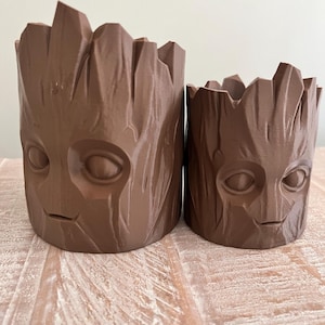 May include: Two brown cylindrical planters with a textured tree bark surface and a sculpted face featuring large oval eyes and a small mouth. The planters are open at the top with jagged, uneven edges resembling broken wood, and they vary in height.