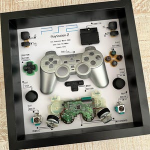 May include: A framed display of a disassembled PlayStation 2 controller. The display includes the shell, face buttons, directional buttons, analog sticks, and motherboard. The text "PlayStation 2" is printed on the white background.