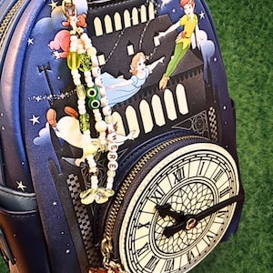 May include: A navy blue backpack featuring a Peter Pan theme with illustrations of Peter Pan, Wendy, and a clock face. The backpack has a gold zipper and a charm with the word "HERE". The clock face shows the time.