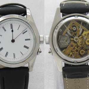 May include: A silver-toned wristwatch with a white face and black Roman numerals. The watch has a black leather band. The back of the watch is transparent, revealing the intricate mechanical workings of the timepiece.