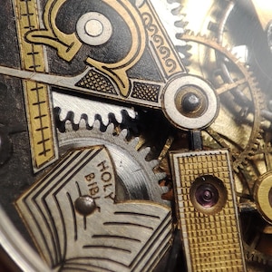 May include: Close-up of a vintage watch mechanism with intricate details. The design features gold and silver accents, gears, and the text "HOLY BIBLE" on a small book. The watch has a complex, layered appearance.