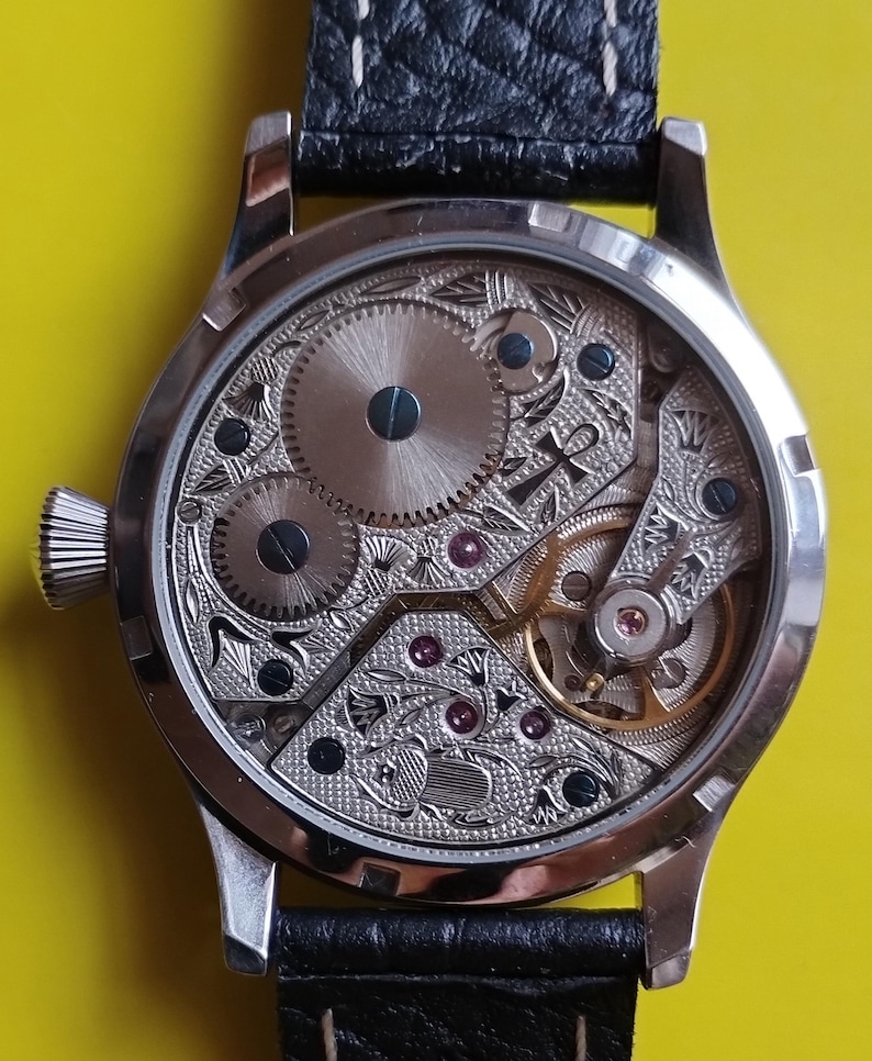 May include: A silver pocket watch with a black leather band. The back of the watch is open, revealing the intricate inner workings of the watch.