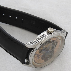 May include: A silver wristwatch with a black leather band. The watch face has a skeleton design with a gold-toned background.