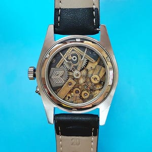 May include: A close-up of a vintage wristwatch with a transparent back, showcasing the intricate mechanical movement. The watch has a gold-toned case, a black leather strap, and a cream-colored band. The watch face displays gears and other internal components.