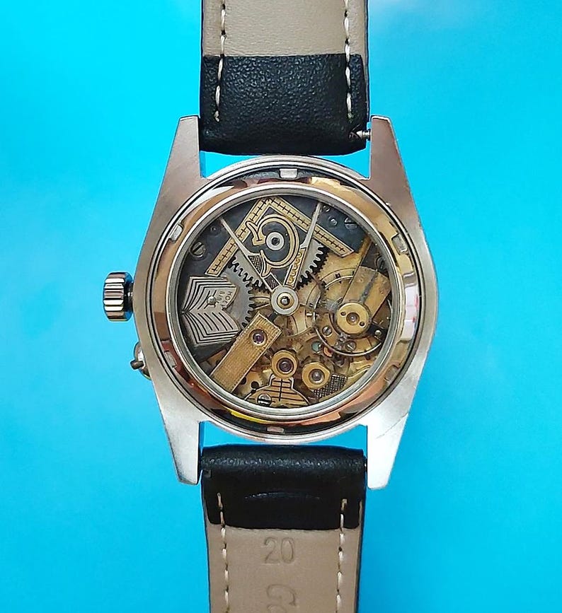 May include: A close-up of a vintage wristwatch with a transparent back, showcasing the intricate mechanical movement. The watch has a gold-toned case, a black leather strap, and a cream-colored band. The watch face displays gears and other internal components.