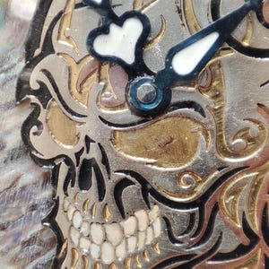 May include: A close-up of a skull-shaped clock face with black and white hands. The skull is made of metal and has a gold and silver finish. The background is a shimmering iridescent material.