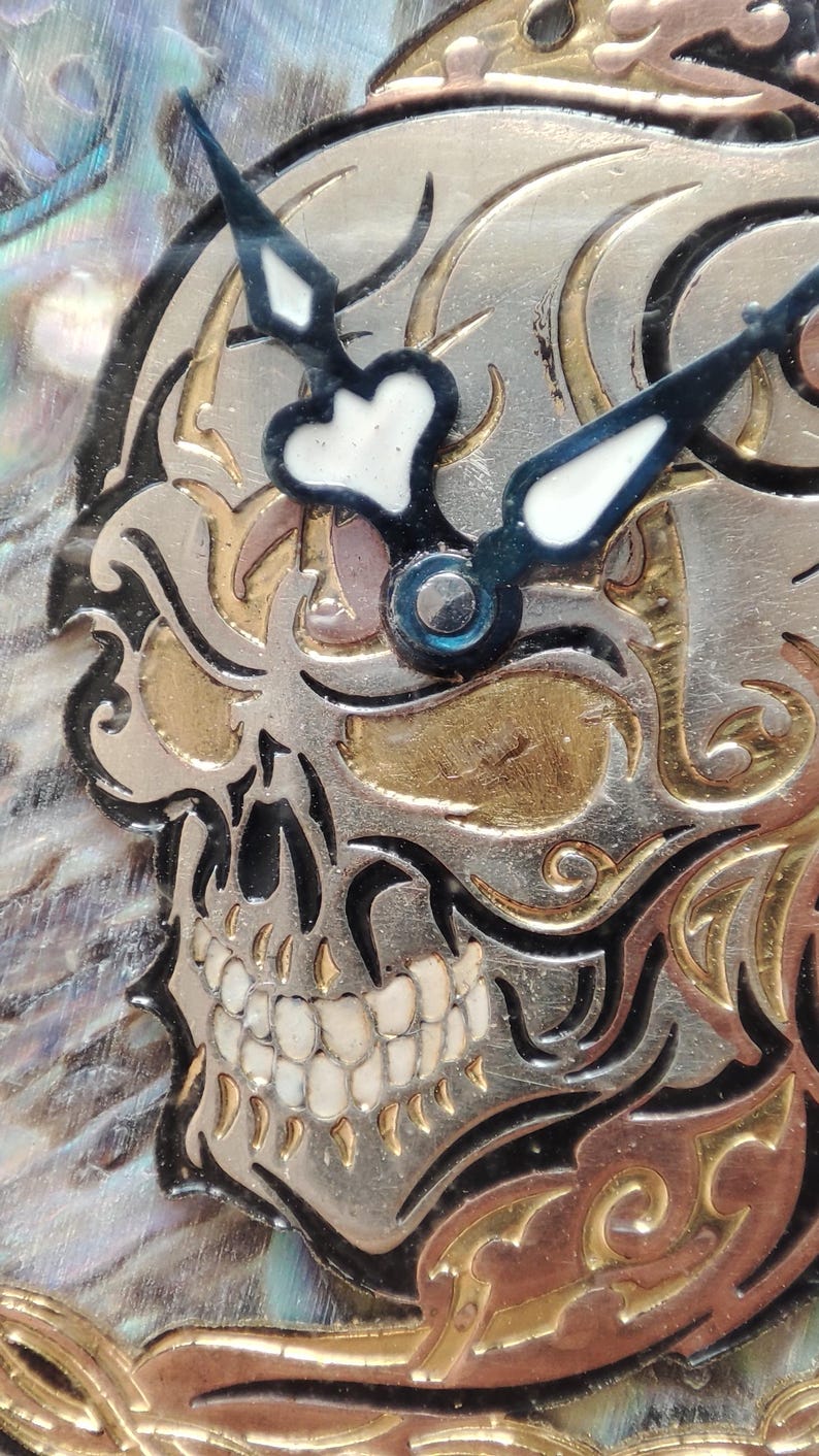 May include: A close-up of a skull-shaped clock face with black and white hands. The skull is made of metal and has a gold and silver finish. The background is a shimmering iridescent material.
