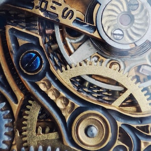 May include: Close-up of a complex watch mechanism with intricate gears and a gold-toned finish. The gears are visible through a clear glass cover, revealing the inner workings of the watch.