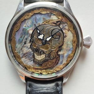 May include: A silver wristwatch with a black leather band. The watch face features a skull design with gold accents and a mother-of-pearl background.