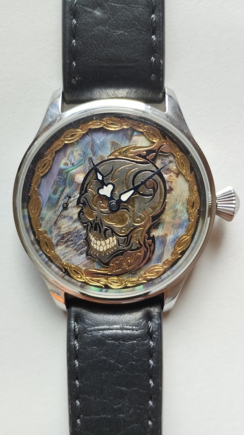May include: A silver wristwatch with a black leather band. The watch face features a skull design with gold accents and a mother-of-pearl background.