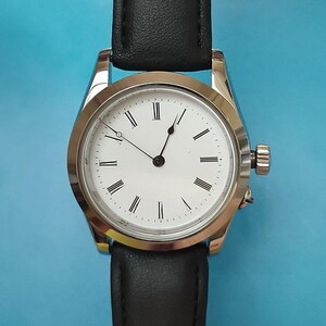 May include: A silver-toned wristwatch with a white face and black Roman numeral hour markers. The watch has black hands and a black leather band. The watch case is round and has a small crown on the side.