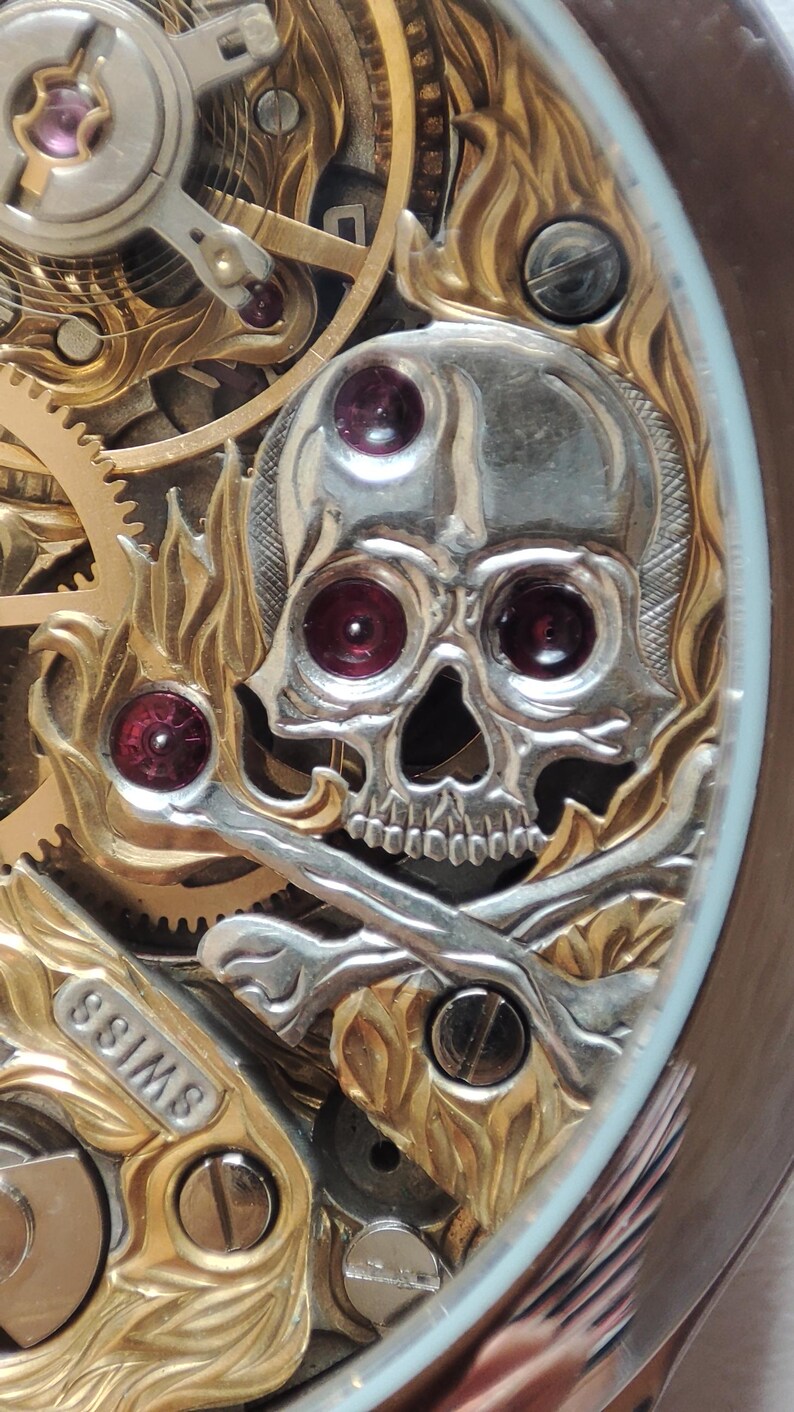 May include: Close-up of a gold and silver watch face with a skull and crossbones design. The skull has red jewels for eyes and the watch face says "Swiss".