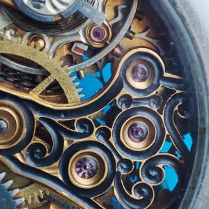 May include: Close-up of a complex, intricate clock mechanism with gold gears and black accents. The mechanism is visible through a clear glass cover.