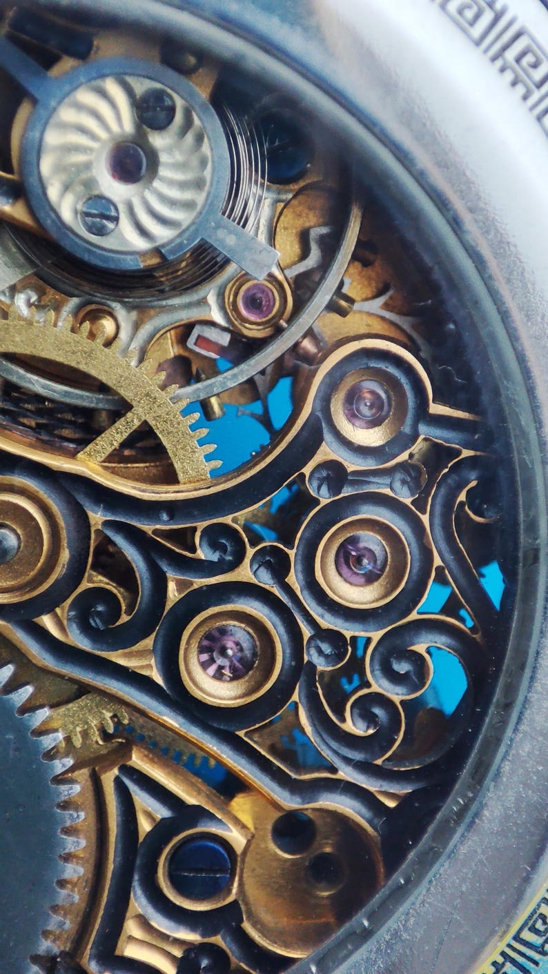 May include: Close-up of a complex, intricate clock mechanism with gold gears and black accents. The mechanism is visible through a clear glass cover.