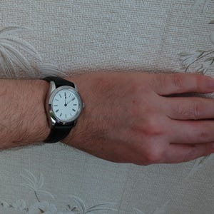 May include: A silver wristwatch with a white face and black hands and markers. The watch has a black leather band. The watch is on a person's wrist.