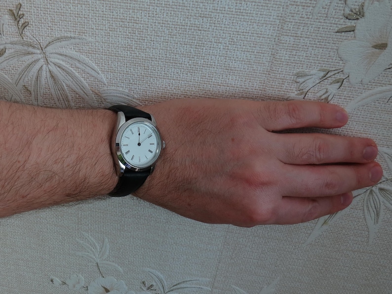 May include: A silver wristwatch with a white face and black hands and markers. The watch has a black leather band. The watch is on a person's wrist.