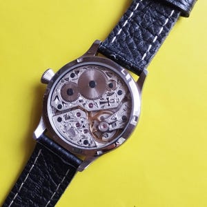 May include: A silver wristwatch with a black leather band and a clear back revealing the inner workings of the watch.