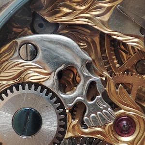 May include: Close-up of a watch mechanism with a gold-toned skull design. The skull is made of metal and is intricately detailed. The watch mechanism is made of metal gears and is visible through a clear glass cover.