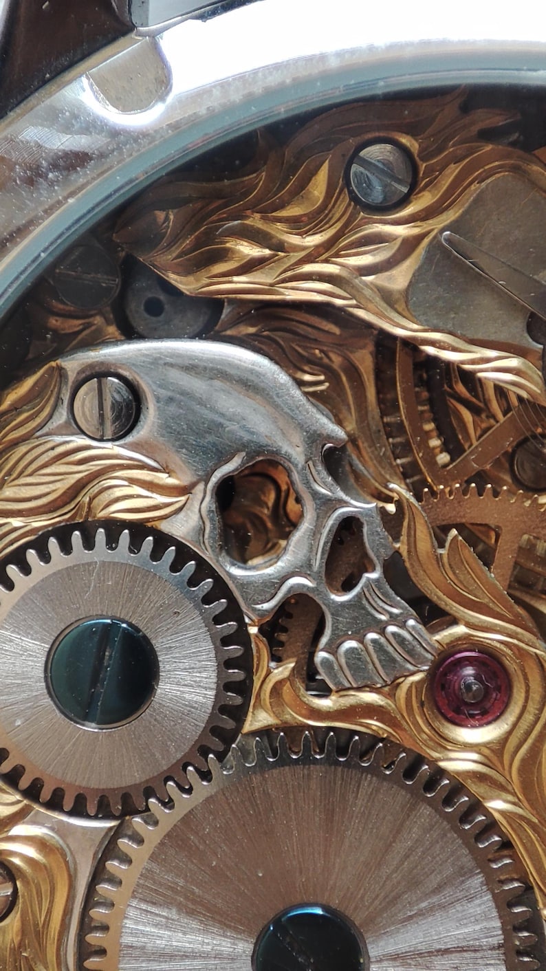 May include: Close-up of a watch mechanism with a gold-toned skull design. The skull is made of metal and is intricately detailed. The watch mechanism is made of metal gears and is visible through a clear glass cover.