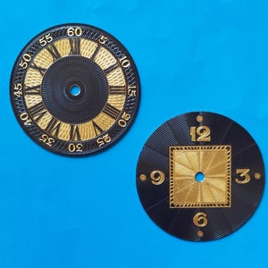 May include: Two circular watch dials with a black and gold colour scheme. The left dial features Roman numerals and a minute track from 5 to 60. The right dial displays a central gold square with a sunburst pattern and the numbers 3, 6, 9, and 12.