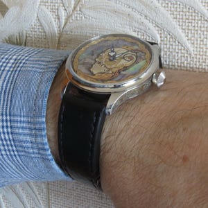 May include: A silver wristwatch with a black leather band. The watch face features a colorful, detailed design.