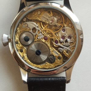 May include: A silver and gold skeleton watch with a black leather band. The watch face is clear and shows the intricate inner workings of the watch. The watch has two skull designs on the face.