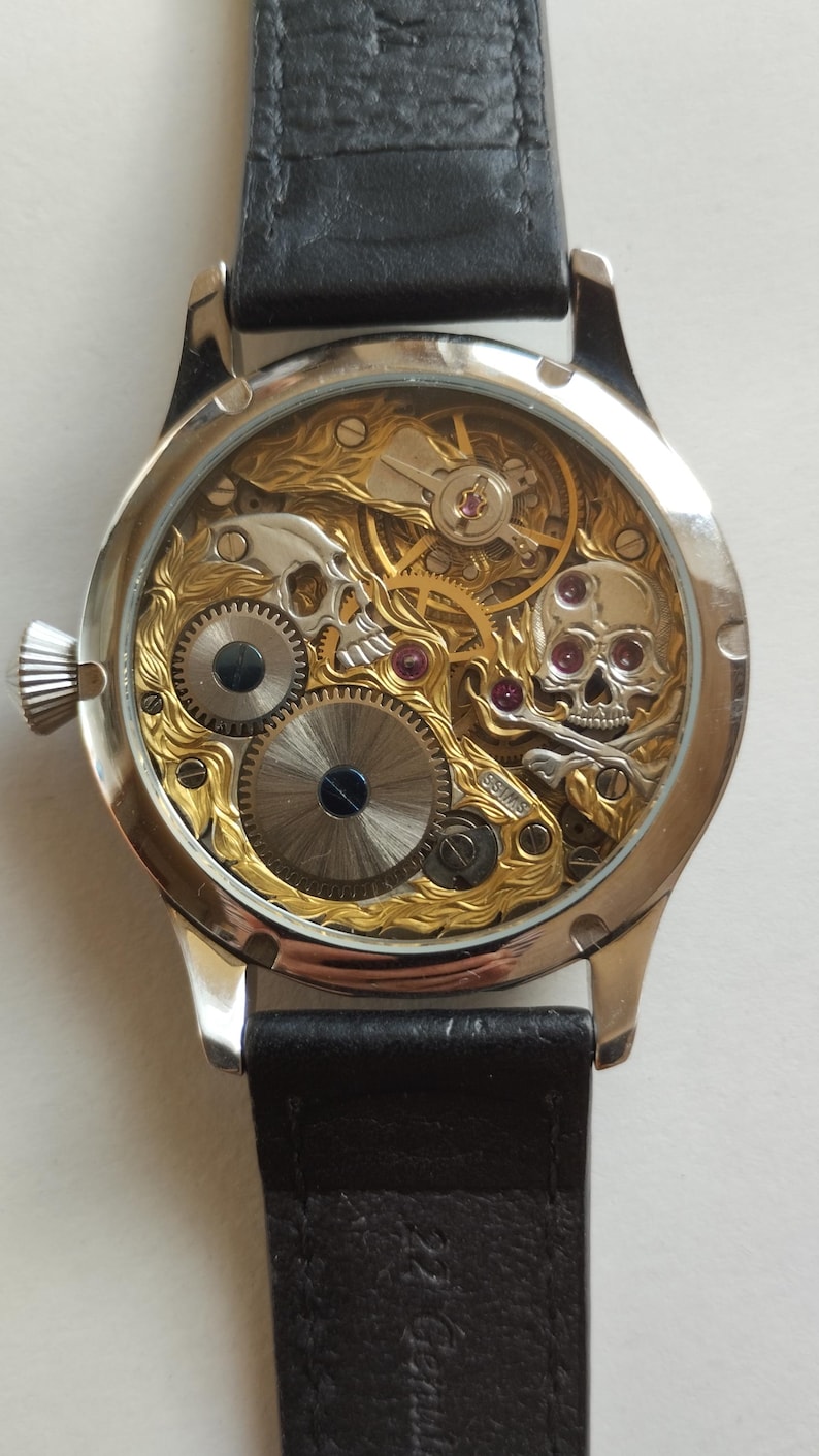 May include: A silver and gold skeleton watch with a black leather band. The watch face is clear and shows the intricate inner workings of the watch. The watch has two skull designs on the face.