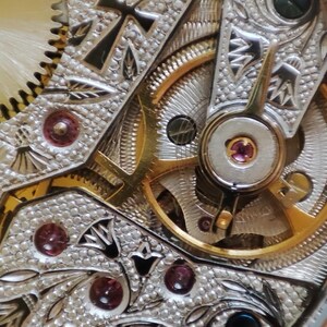 May include: Close-up of a detailed, intricate watch mechanism with gold gears and a silver-toned base. The mechanism features an ankh symbol and decorative patterns.