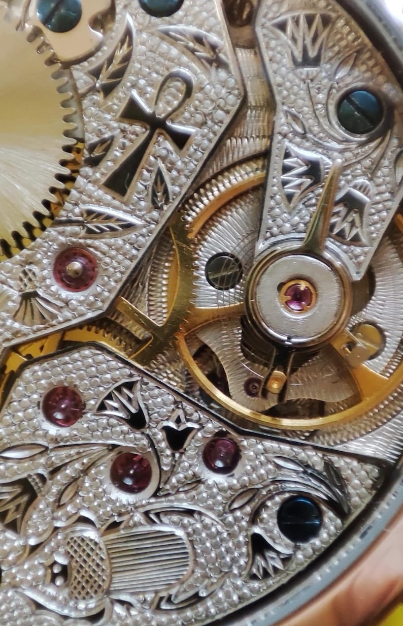 May include: Close-up of a detailed, intricate watch mechanism with gold gears and a silver-toned base. The mechanism features an ankh symbol and decorative patterns.