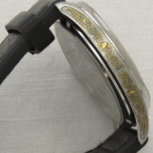 May include: A silver watch with a black leather band. The watch face is surrounded by a gold-colored band with an intricate design.