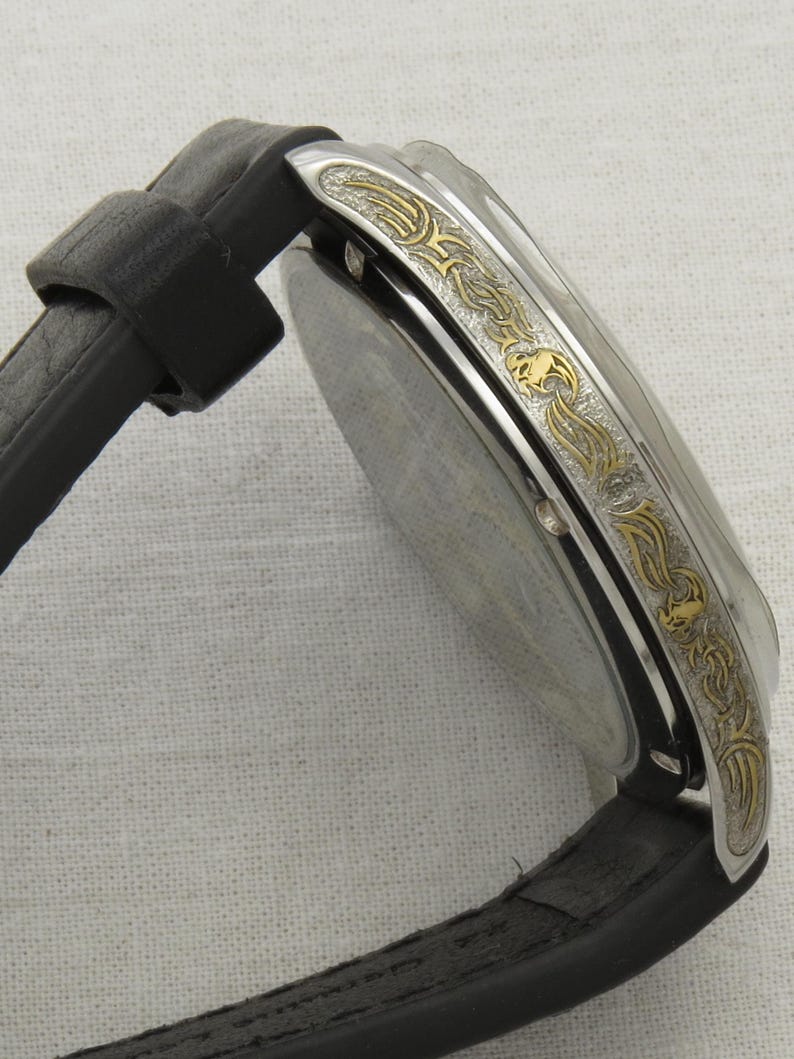 May include: A silver watch with a black leather band. The watch face is surrounded by a gold-colored band with an intricate design.